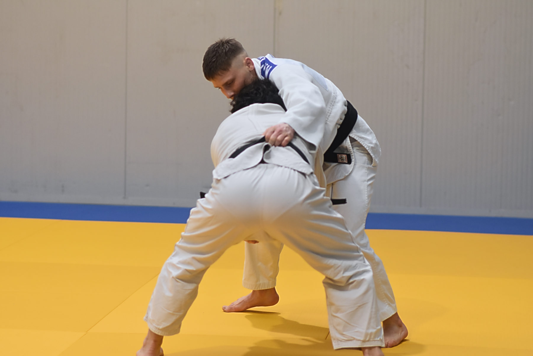 Due judoka in una presa in piedi sul tatami giallo, con la parete grigia dietro