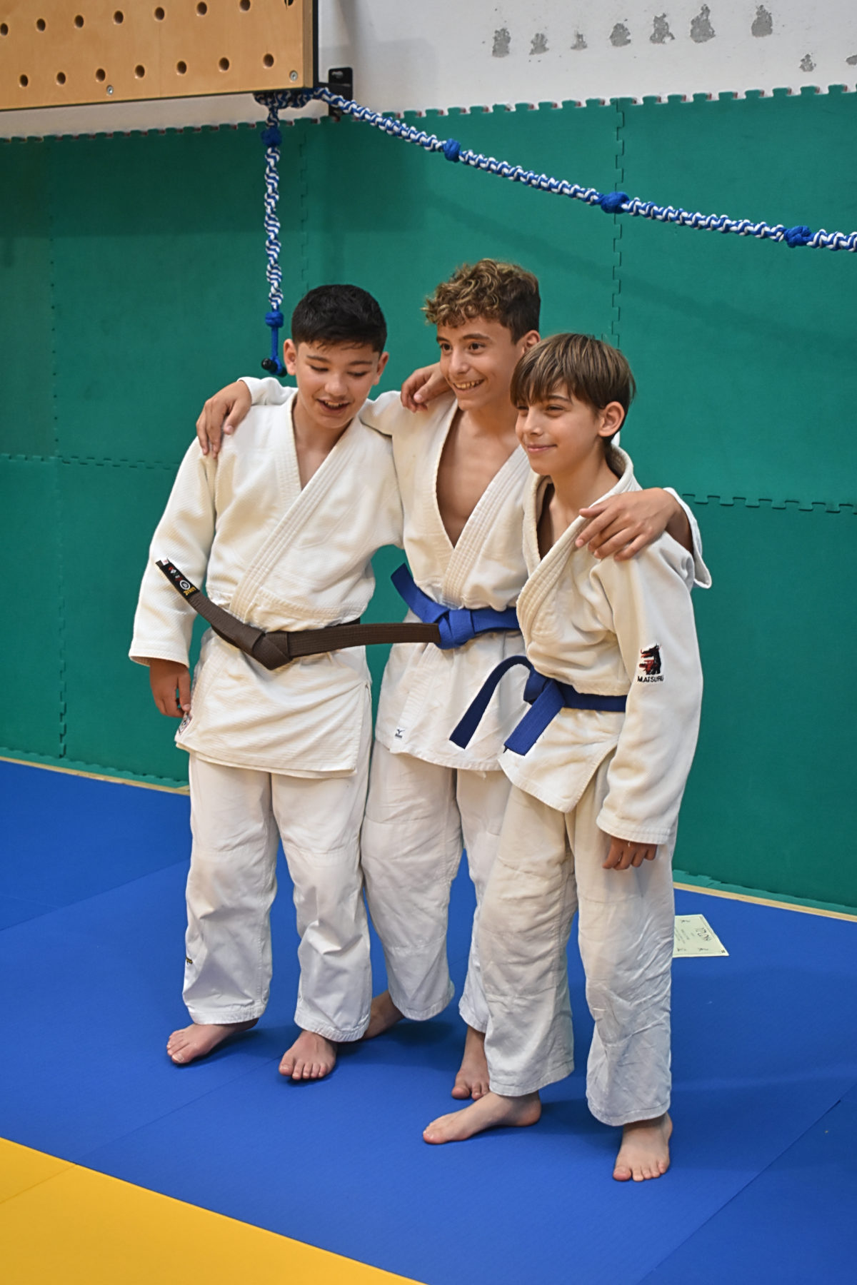 Tre ragazzi in judogi abbracciati sorridono davanti a un muro verde